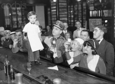 29 Dec 1931, Havana, Cuba --- 12/29/31-Havana, Cuba: Sloppy Joe, Jr., just four years of age and having two years behind the bar, is celebrating his graduation from apprenticeship by mixing his real champagne cocktail behind his father's world-famed bar. Sloppy Joe Jr., is quite proficient at mixing the more common varieties. Here, customers toast the little guy. --- Image by © Bettmann/CORBIS